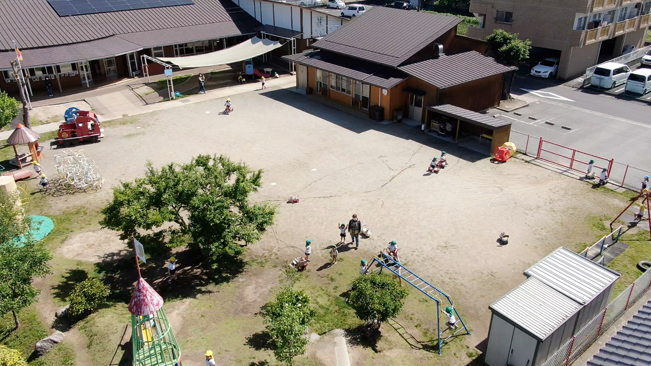 広い園庭と木造平屋の園舎。木かげや遊具のまわりで子どもたちが遊んでいます