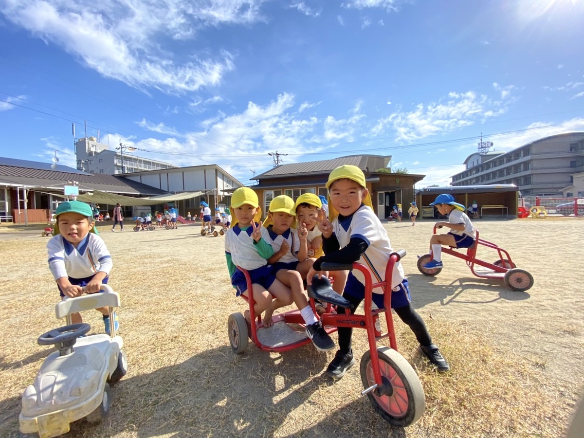 青空の下、園庭で大きな乗り物をみんなで動かす子どもたち