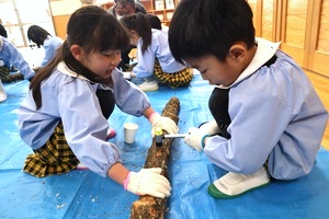 床にひろげたシートの上で、軍手をはめて丸太の皮をけずる二人の子ども