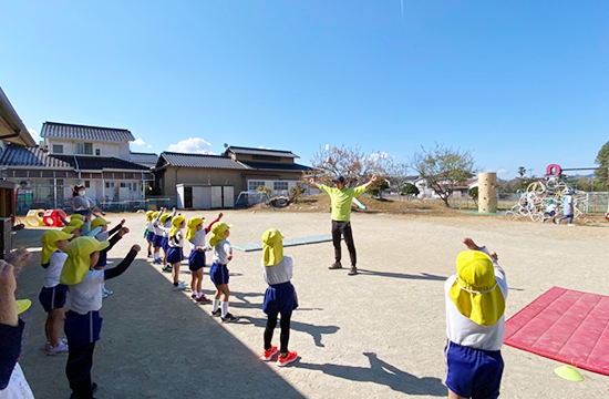 桜の咲く園庭で、先生を囲んで並び、順番を待つ子どもたち