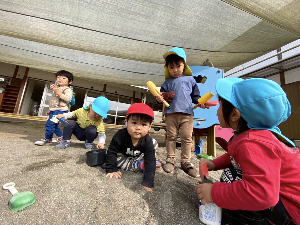 砂場のそばでしゃがみこみ、道具を手に遊ぶ子どもたち