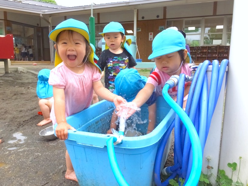 青いたらいに張った水を、ホースの水といっしょに手ですくってあそぶ小さな子どもたち