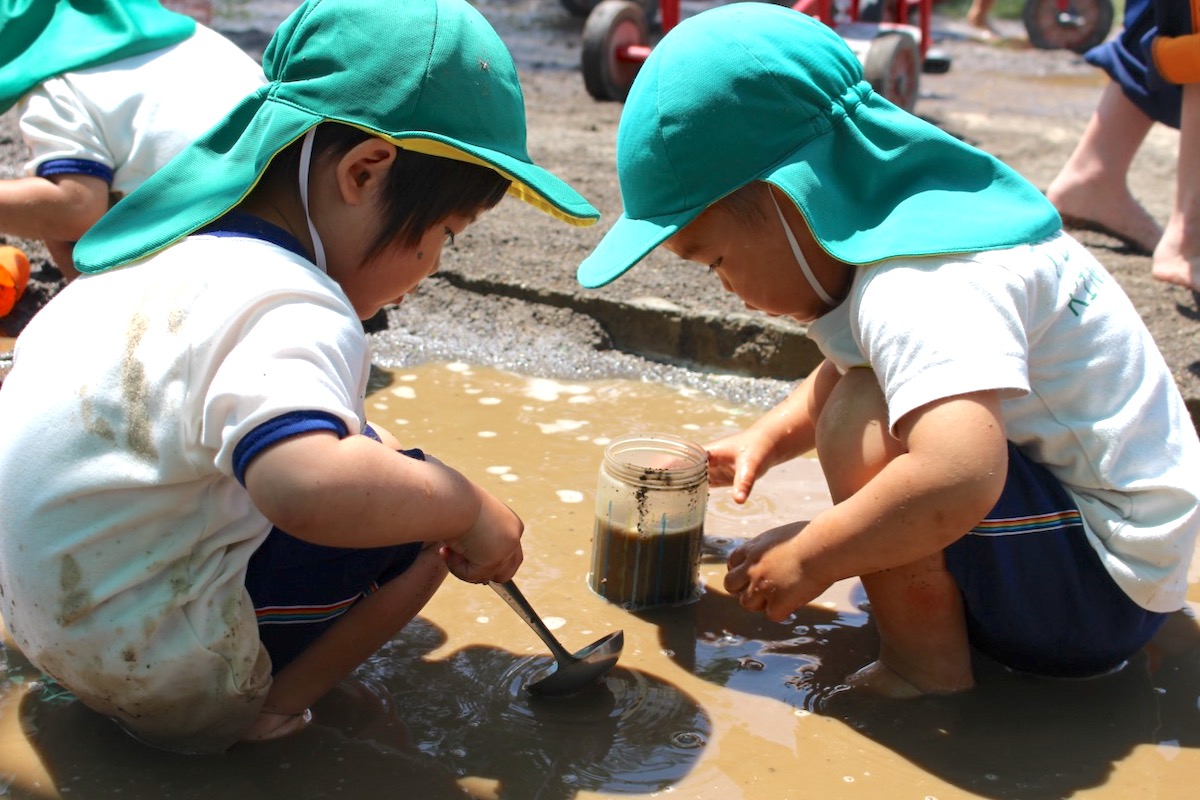 雨あがりの水たまりのそばで、びんとおたまを使って泥水をすくって試している二人の子ども