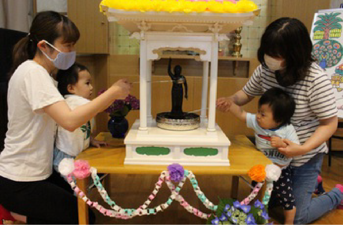 花御堂のみほとけさまに、保育者に抱かれて手をのばす小さな子どもたち