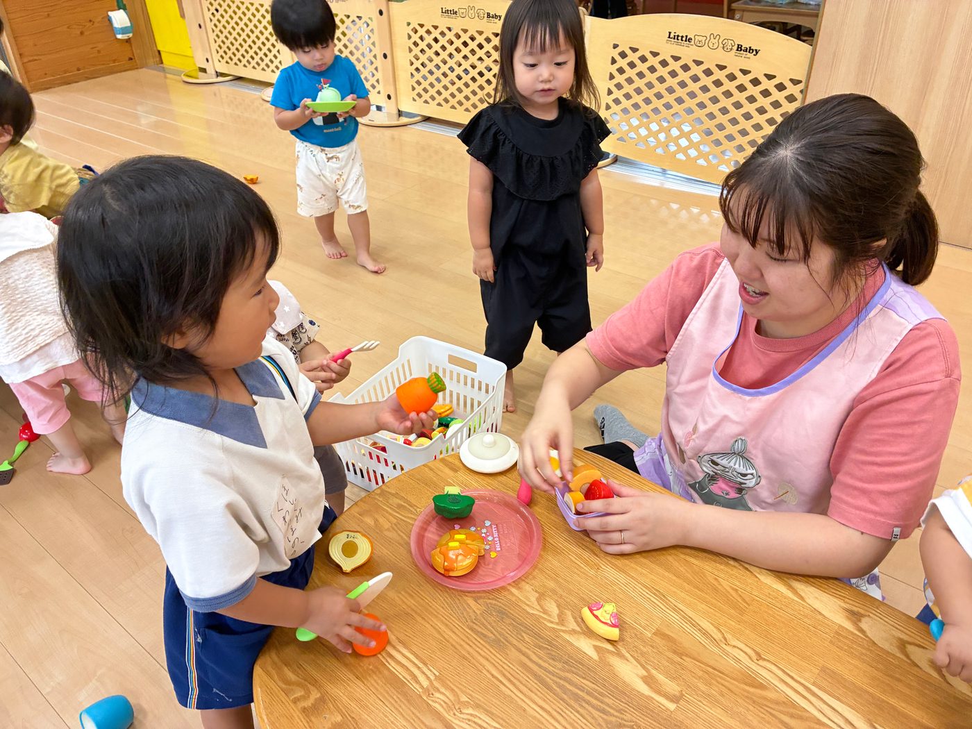 保育室で、ままごとの道具を手に、子どもと目線を合わせて遊ぶ保育者