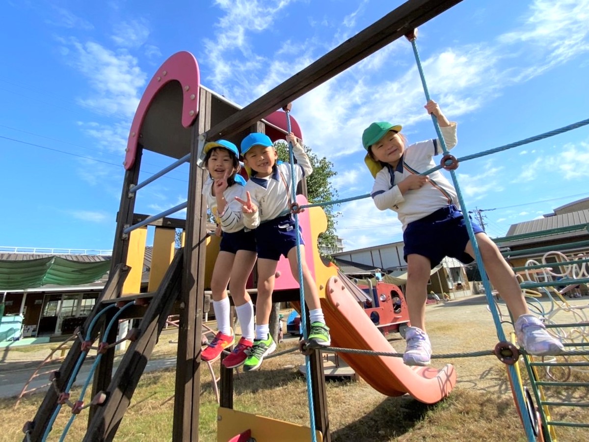 青空の下、園庭の遊具によじ登って笑い合う子どもたち